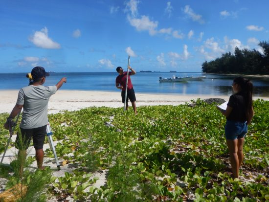 The background is a vegetated sandy beach north of Sugar Dock, where a boat is docked. On the left is Ross Arriola pointing next to the mounted Berger Level. Thomas Benavente is holding the rod level in the middle. Kiana is looking towards Thomas with the record book in hand.