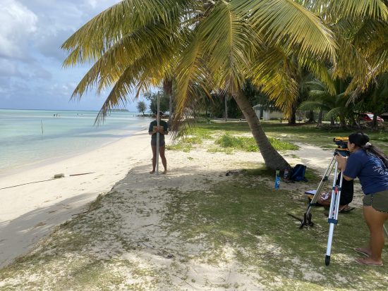 At a beach between Crowne Plaza and Hyatt, Kyla looks through the Berger level, an instrument mounted on a tripod. Emma holds a rod level, a measuring instrument for height, underneath a coconut tree. The beach looks damaged, but the ocean is clear, shallow, and calm during low tide conditions.